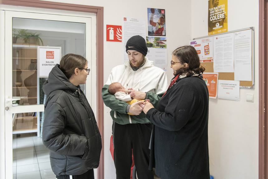 Un papa tient son bébé dans les bras. A côté de lui se trouve la maman et une assitante sociale.