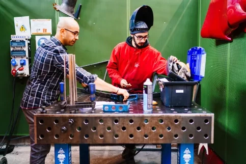 Un jeune homme porte des protections et s'apprête à faire de la soudure. Il est aidé par un professeur.