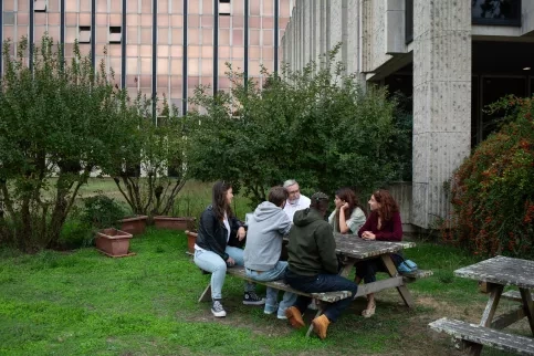 Un groupe 6 personnes discutent autour d'une table, ils se trouvent devant un grand immeuble, sur une table en bois.
