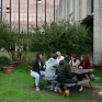 Un groupe 6 personnes discutent autour d'une table, ils se trouvent devant un grand immeuble, sur une table en bois.